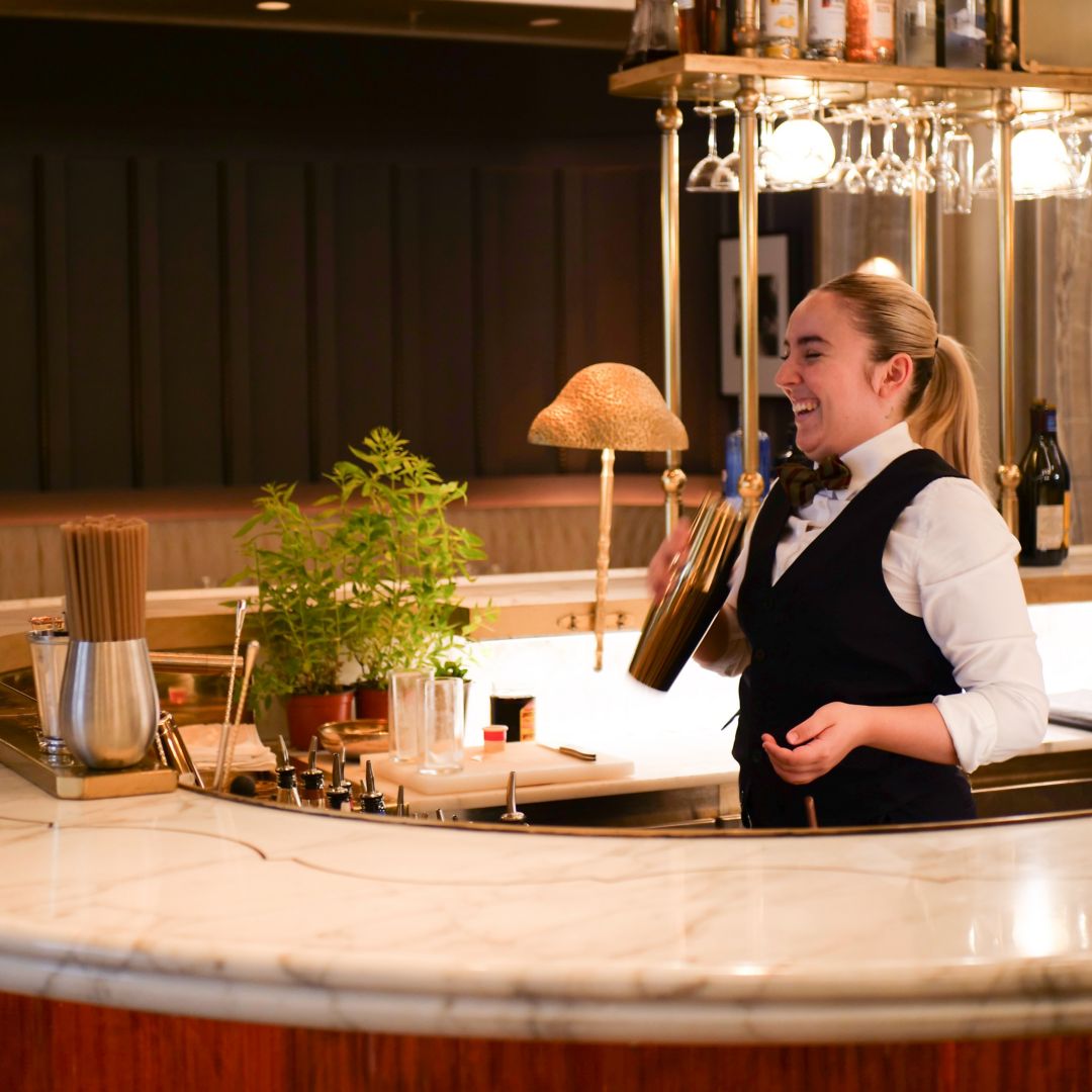 A bartender in a white shirt and black vest smiling as she skillfully shakes a cocktail shaker.